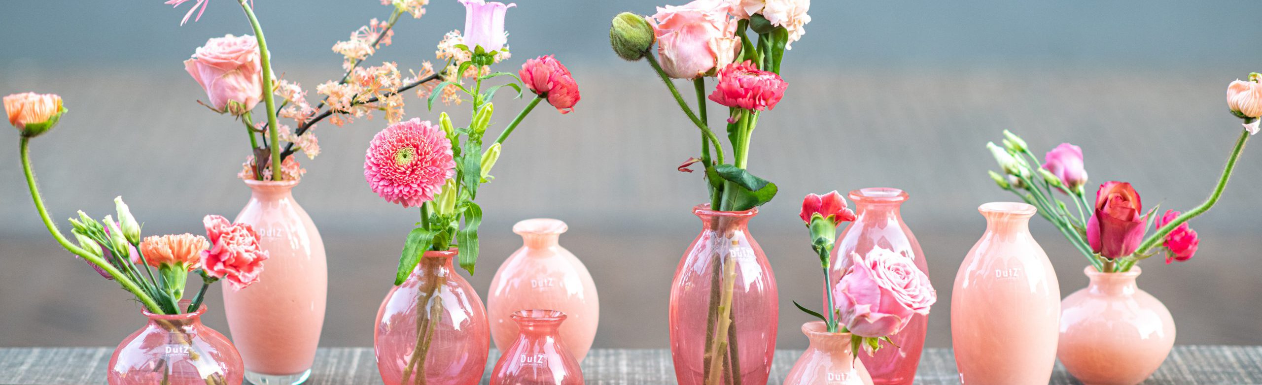 DutZ_nadiel_apricot_3 DutZ vaasjes uit de serie Nadiel in verschillende roze tinten. Alle vaasjes zijn gevuld met roze bloemen. De vaasjes staan op een rij naast elkaar op een tafel