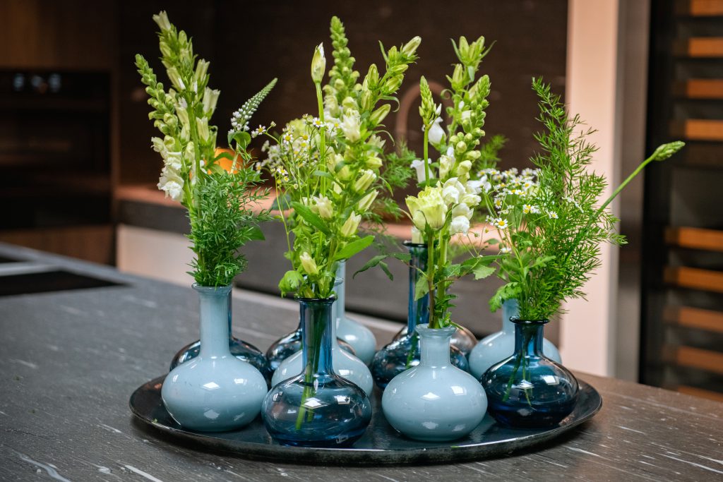 Blauwe vaasjes, transparant en opaque, varierend in hoogte en gevuld met bloemen op een glazen onderbord
