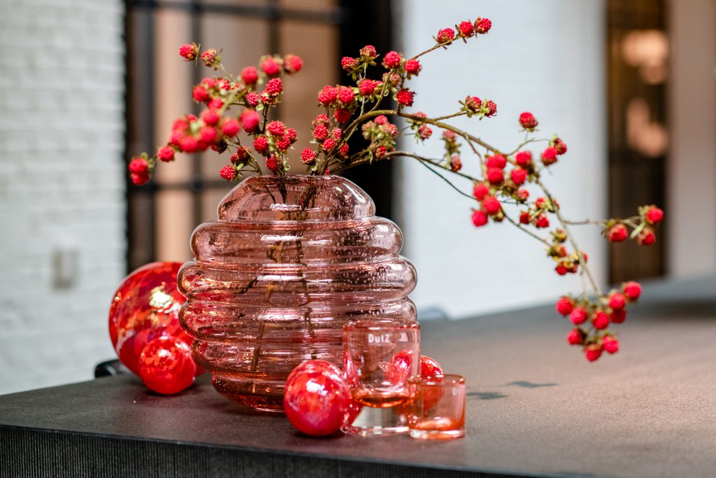 Roze Honey Barrel vaas op een tafel. Voor deze vaas liggen rode decoratieve ballen. Witte muur op de achtergrond