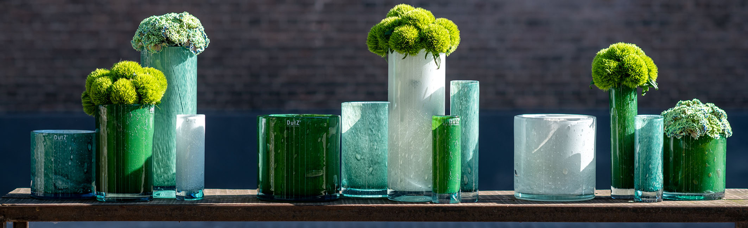 DutZ gerecyclede vazen in groen en wittinten in een rij op een buitentafel. De vazen varieren in formaat en enekel zijn gevuld met bloemen